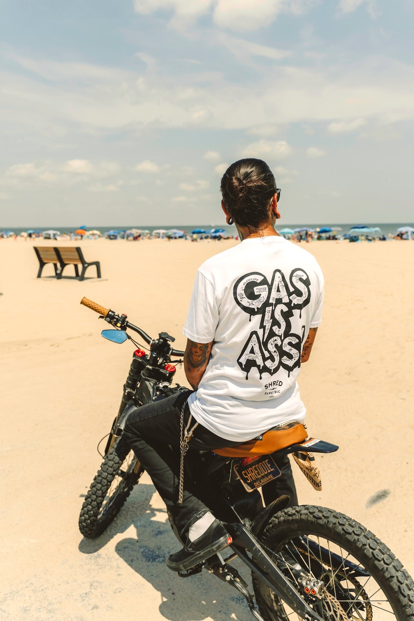 Person riding an electric bike with 'Gas Is Ass' anti-gas shirt on the beach