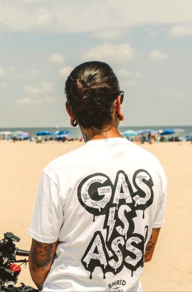 Person wearing a white t-shirt with Gas Is Ass text on the beach