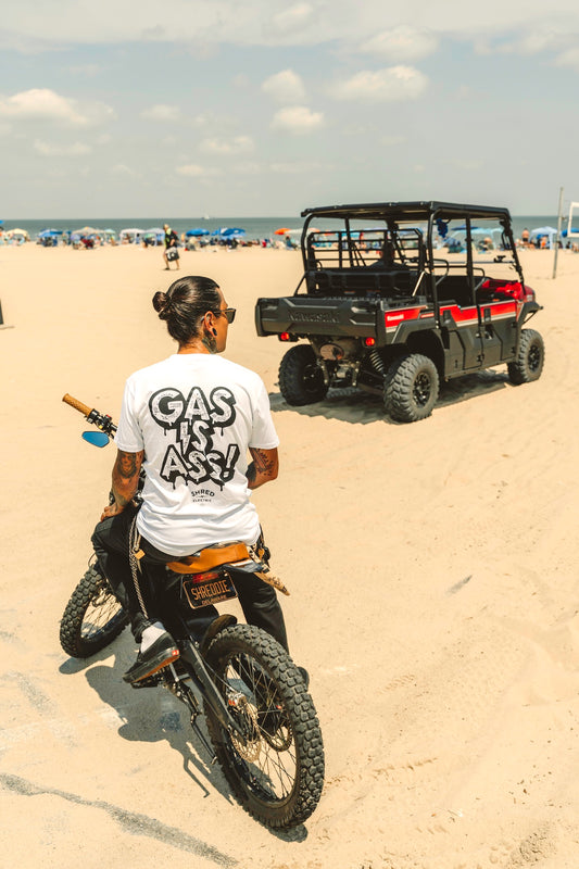 Person wearing Gas Is Ass! Shirt on beach with an electric dirt bike and off road vehicle