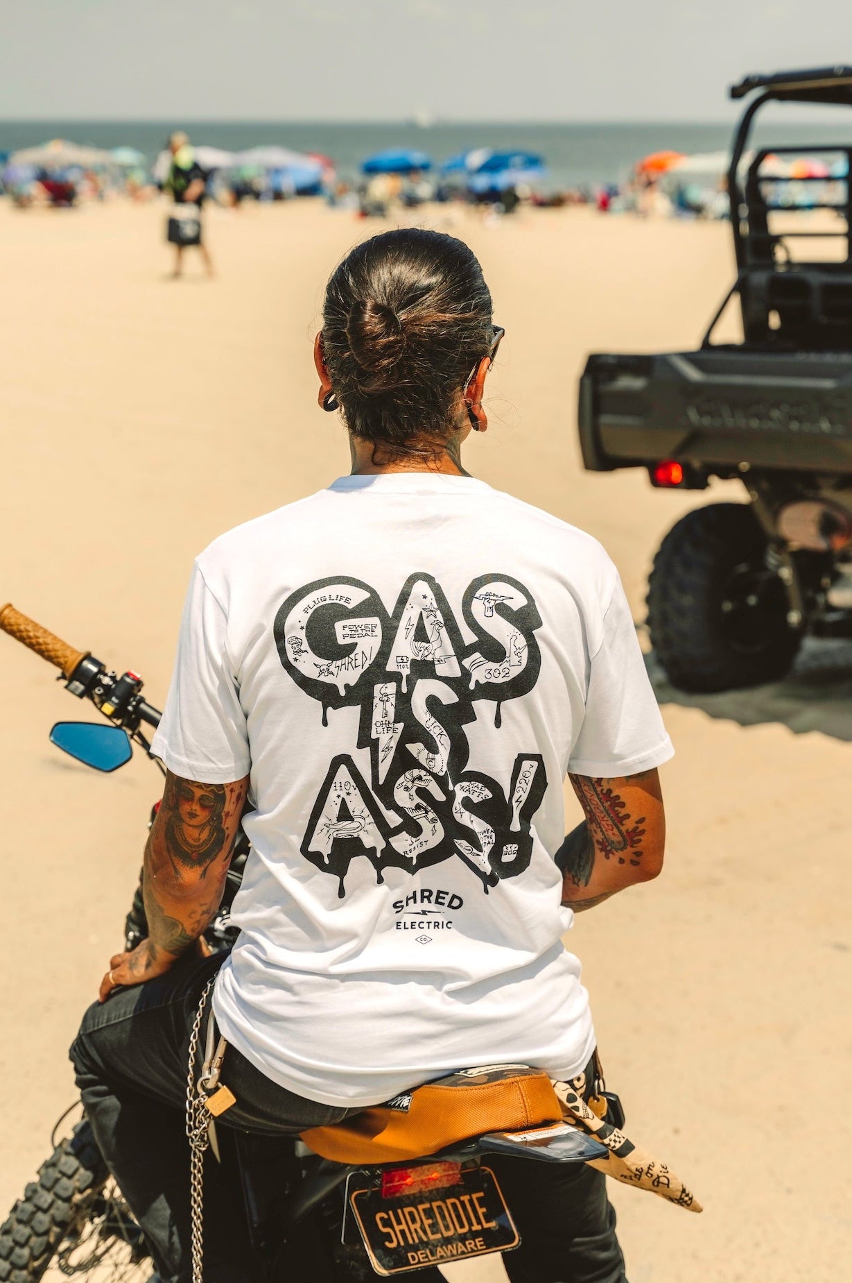 Gas Is Ass white tattoo print electric t-shirt back view on a bike on the beach
