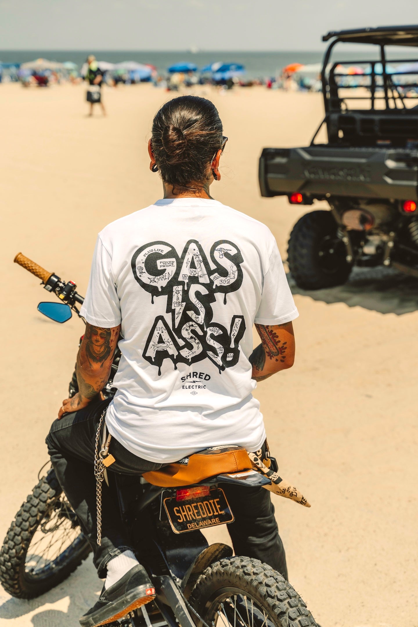 'GAS IS ASS!' white t-shirt with an electric vehicle and beach in background.