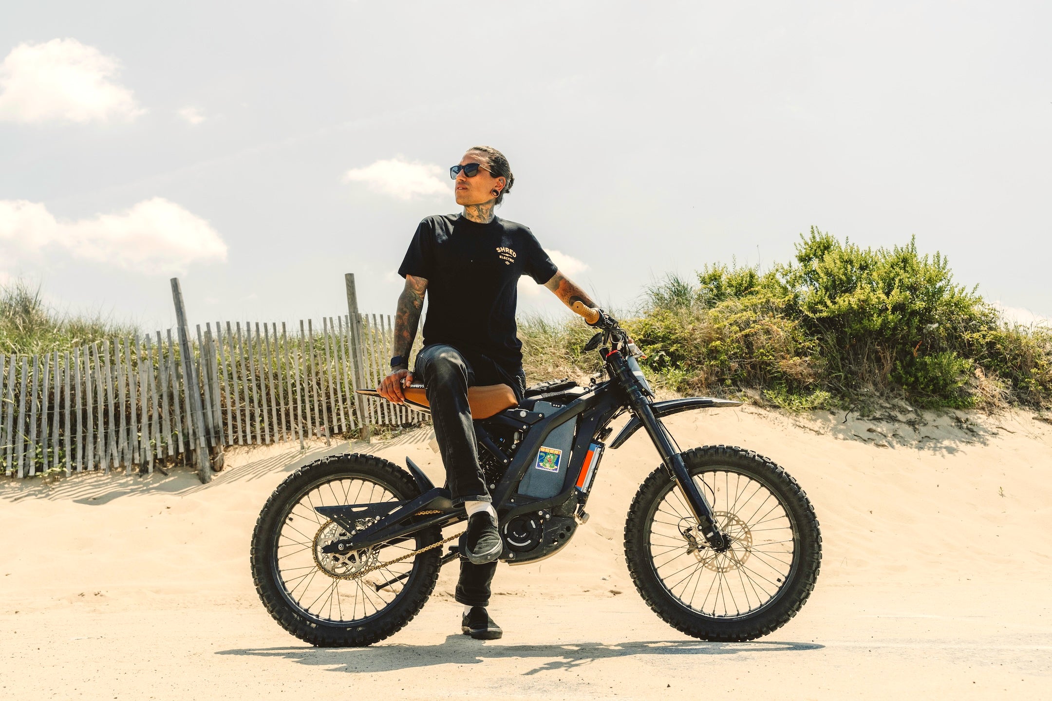 Person riding a Sur Ron electric dirt bike on a sandy beach with sky in the background