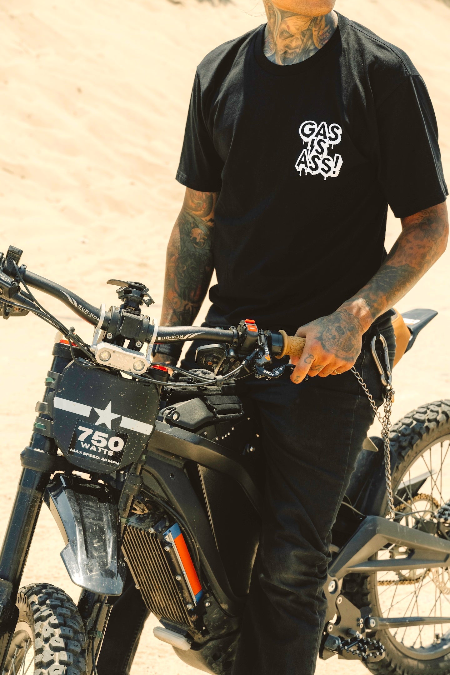 Person wearing a black t-shirt with a logo, standing next to an electric motorcycle in a desert setting.