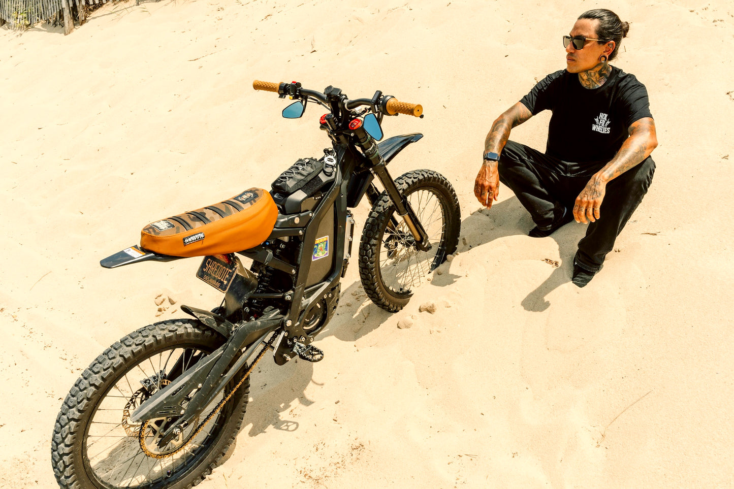 Honk For Wheelies black shirt front view on person sitting on sand next to an electric Dirtbike