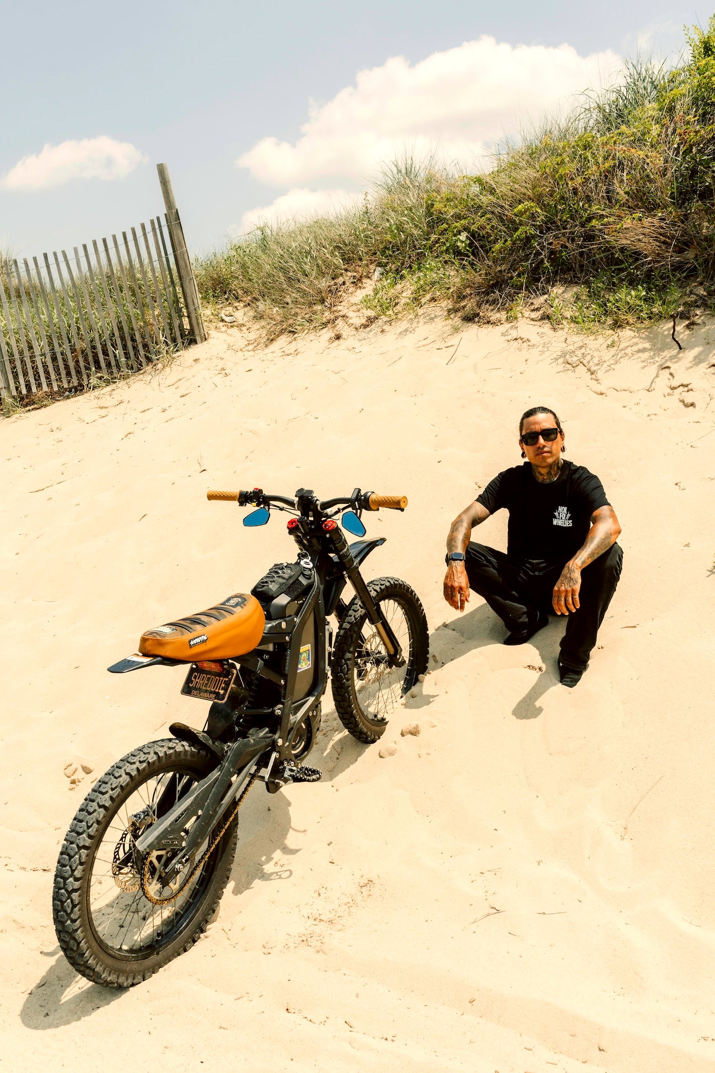 Person sitting on a sand dune with a motorcycle, wearing sunglasses and a black 'honk for wheelies' shirt.