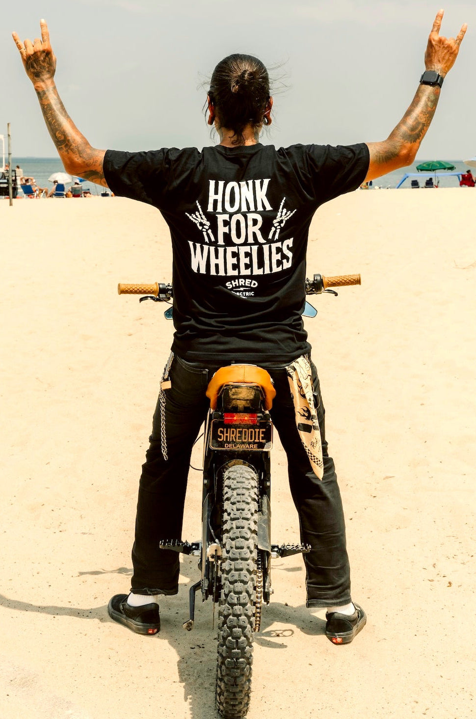 Person on a dirt bike wearing a black t-shirt with 'Honk for Wheelies' text, standing on a sandy beach.