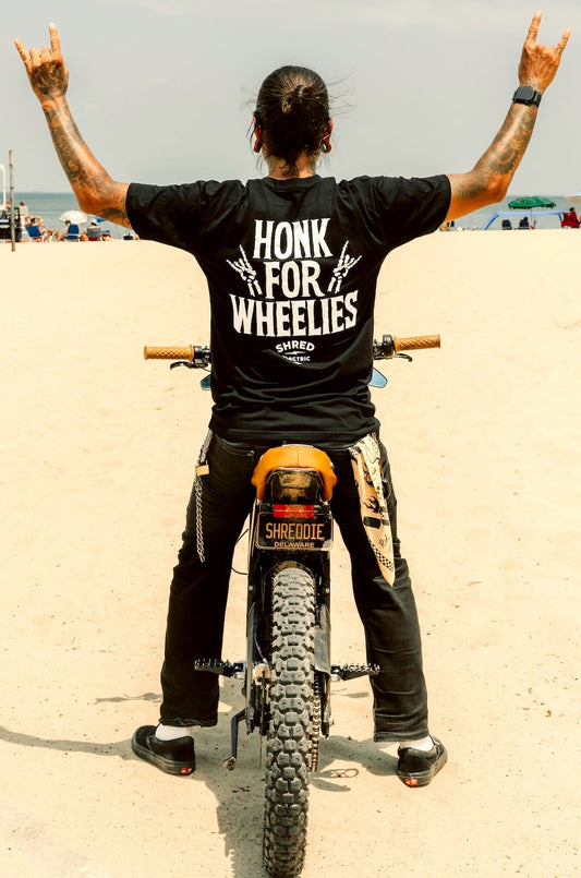 Person on a dirt bike wearing a black t-shirt with 'Honk for Wheelies' text, standing on a sandy beach.