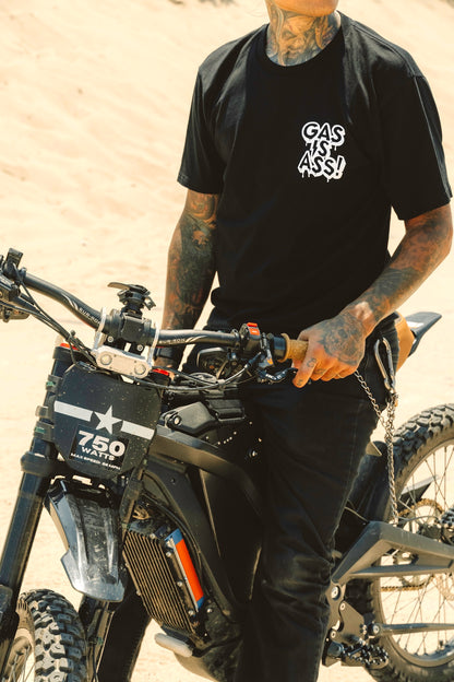 Person wearing a black t-shirt with a logo, standing next to an electric motorcycle in a desert setting.