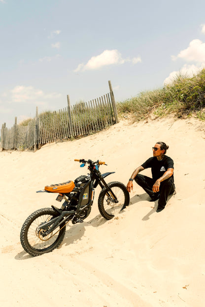 Honk for wheelies black t-shirt on person sitting next to a bike on a sandy beach 
