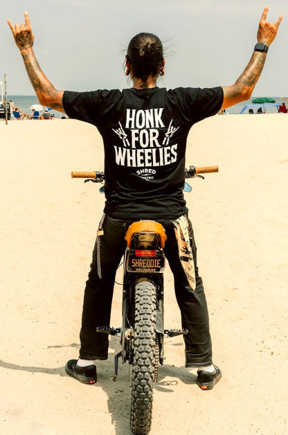 Person on a dirt bike wearing a black t-shirt with 'Honk for Wheelies' text, standing on a sandy beach.